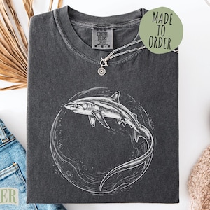 May include: Dark gray t-shirt with a white shark illustration inside a circle. The shirt has a crew neck and short sleeves. A silver necklace with a small pendant is on the shirt. The words "MADE TO ORDER" are on a green circle.
