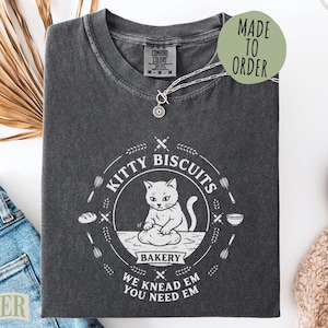 May include: A charcoal gray t-shirt featuring a white graphic of a cat kneading dough. The circular design includes the text "KITTY BISCUITS BAKERY" and "WE KNEAD EM YOU NEED EM" surrounded by illustrations of rolling pins, whisks, and bread.