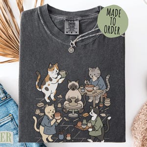May include: A charcoal gray short-sleeve t-shirt featuring a graphic illustration of five cats working in a pottery studio. The cats are shown shaping clay on a wheel, painting, and holding ceramic mugs. The shirt has a crew neckline and a tag.