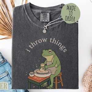 May include: A charcoal gray t-shirt featuring a graphic of a green frog wearing an apron and working at a pottery wheel. The text "i throw things" is printed in a light color above the frog. The shirt has a crew neckline and a tag.