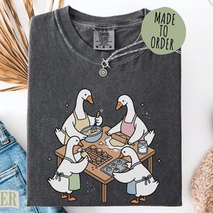 May include: A charcoal gray t-shirt featuring a graphic of four white geese wearing aprons and baking at a wooden table. The geese are shown whisking batter, rolling dough, piping frosting, and placing cookies on a tray. The shirt has a crew neckline.