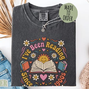 May include: A charcoal gray t-shirt featuring a graphic print with the text "I've Been Reading Since 1900s". The design includes a smiling sun, an open book, colorful flowers, and hearts in shades of pink, yellow, blue, and orange.