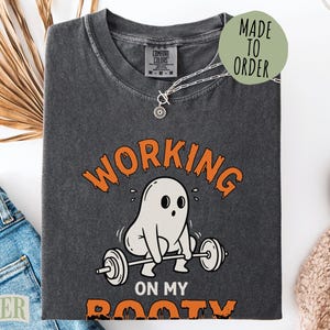 May include: A charcoal gray t-shirt featuring a graphic of a white ghost lifting a barbell. The text reads "WORKING ON MY BOOTY" in orange letters. A circular green tag on the shirt says "MADE TO ORDER". The shirt has a crew neck and short sleeves.