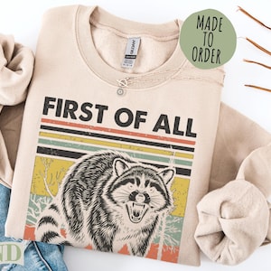 Crewneck sweatshirt with "FIRST OF ALL" and a raccoon graphic.