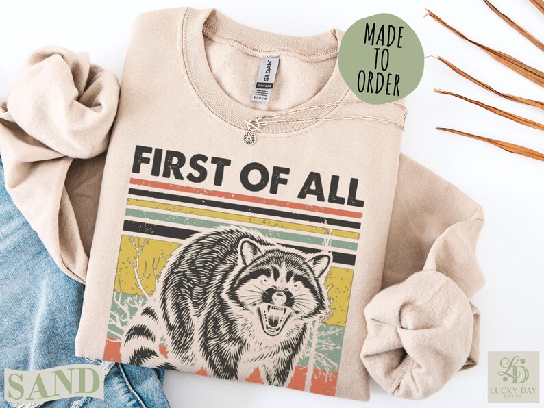 Crewneck sweatshirt with "FIRST OF ALL" and a raccoon graphic.