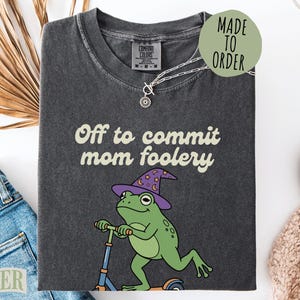 May include: Dark gray t-shirt with the phrase "Off to commit mom foolery" and a cartoon frog wearing a witch hat and riding a scooter. The shirt has a Comfort Colors tag and a Made to Order sticker.