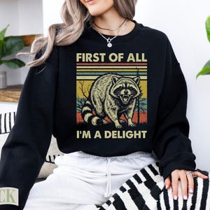 Black crewneck sweatshirt with raccoon and text "First of all I'm a delight".