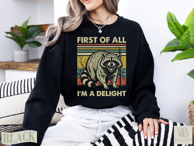 Black crewneck sweatshirt with raccoon and text "First of all I'm a delight".