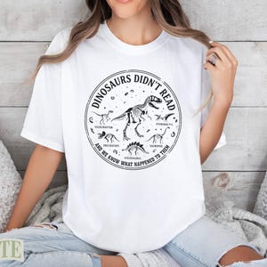 White t-shirt with black circular graphic featuring dinosaurs and text about them not reading.