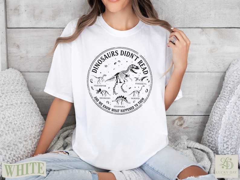 White t-shirt with black circular graphic featuring dinosaurs and text about them not reading.