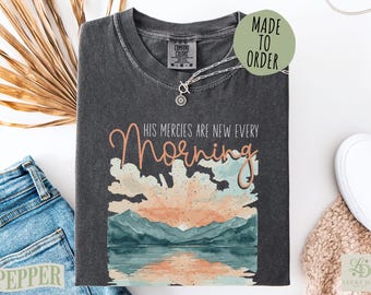 Może przedstawiać: Ciemnoszary t-shirt z grafiką przedstawiającą wschód słońca nad górami i jeziorem. Napis "HIS MERCIES ARE NEW EVERY Morning" wykonano czcionką łączącą styl drukowany i pisany. Koszulka posiada okrągły dekolt oraz metkę z napisem Comfort Colors.