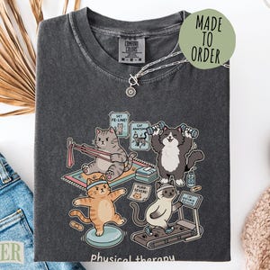 May include: A charcoal gray t-shirt featuring a graphic of four cats performing physical therapy exercises. The design includes cats using resistance bands, dumbbells, a balance board, and a treadmill, with text labels including Get Fe-Line and Cat Anatomy.
