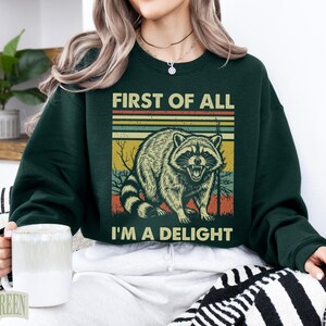 Dark green sweatshirt with raccoon and "First of all, I'm a delight" text.