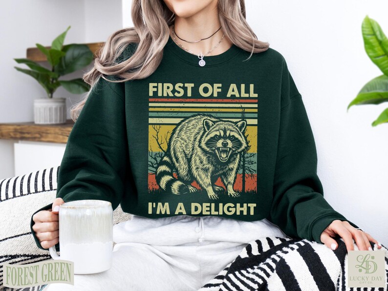 Dark green sweatshirt with raccoon and "First of all, I'm a delight" text.