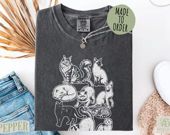 Puede incluir: Una camiseta de manga corta gris carbón con un estampado gráfico blanco de varias razas de gatos en diferentes posturas. La camiseta tiene cuello redondo y una etiqueta de la marca Comfort Colors en el interior. Una etiqueta verde dice MADE TO ORDER.