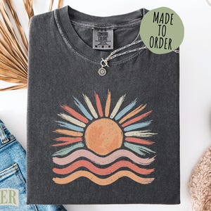 May include: A charcoal gray short-sleeve t-shirt featuring a graphic of a sun rising over waves. The sun is orange with multi-colored brushstroke rays in blue, red, and cream. The waves below are depicted in wavy lines of pink, red, and orange.