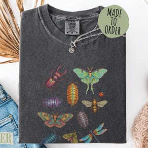 May include: Dark gray Comfort Colors t-shirt featuring colorful insect illustrations. The shirt includes a variety of bugs, including a beetle, moth, bee, and dragonfly. A silver necklace with a small pendant is also visible. The shirt has the text "MADE TO ORDER".