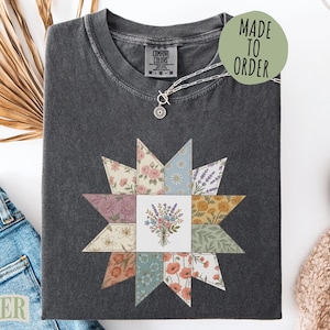 May include: A charcoal gray short-sleeve t-shirt featuring a central square graphic of a flower bouquet surrounded by a star-shaped quilt pattern composed of various floral and botanical prints in muted tones of pink, blue, yellow, and green.