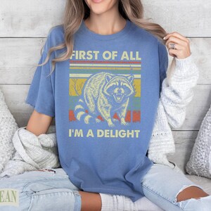Blue t-shirt with vintage raccoon graphic and text "First of all I'm a delight."
