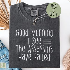 May include: Dark gray t-shirt with the text "Good Morning I See The Assassins Have Failed" in white. The shirt is a Comfort Colors brand and is made to order. A silver necklace with a pendant is on the shirt.