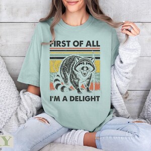 Raccoon graphic with text "FIRST OF ALL I'M A DELIGHT" on a light green t-shirt.