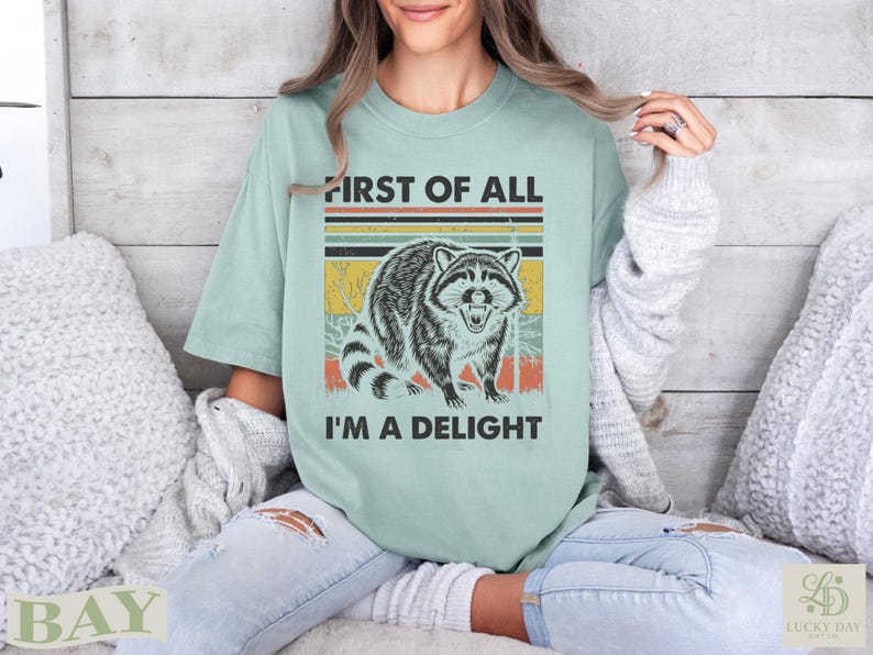 Raccoon graphic with text "FIRST OF ALL I'M A DELIGHT" on a light green t-shirt.