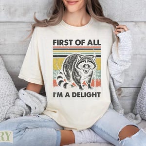 Cream t-shirt with raccoon graphic and text "FIRST OF ALL I'M A DELIGHT".