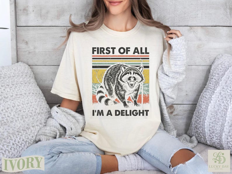 Cream t-shirt with raccoon graphic and text "FIRST OF ALL I'M A DELIGHT".