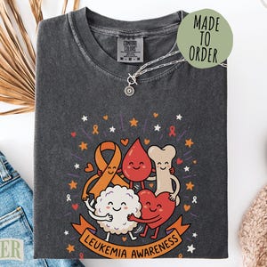 May include: A charcoal gray t-shirt featuring a graphic of cartoon characters representing leukemia awareness, including an orange ribbon, a blood drop, a bone, and a heart. The text "LEUKEMIA AWARENESS" is printed on an orange banner at the bottom.