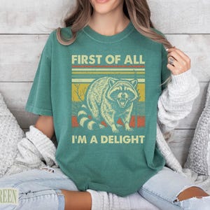Green t-shirt with raccoon and "First of all I'm a delight" text.
