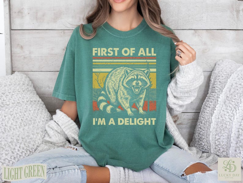 Green t-shirt with raccoon and "First of all I'm a delight" text.