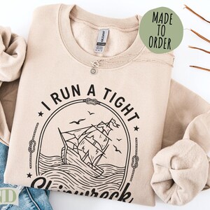 Sand-colored "I Run a Tight Ship" graphic sweatshirt with a sailing ship illustration.