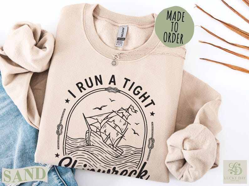 Sand-colored "I Run a Tight Ship" graphic sweatshirt with a sailing ship illustration.