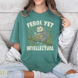 Feral Yet Intellectual text on a green t-shirt featuring an opossum reading a book.