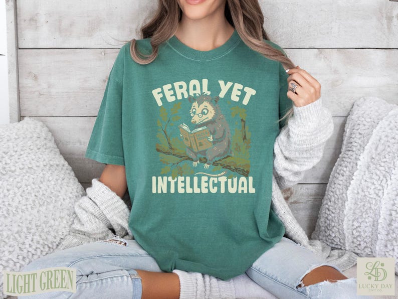 Feral Yet Intellectual text on a green t-shirt featuring an opossum reading a book.