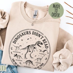 Sand sweatshirt with "Dinosaurs Didn't Read" graphic and dinosaur skeletons.