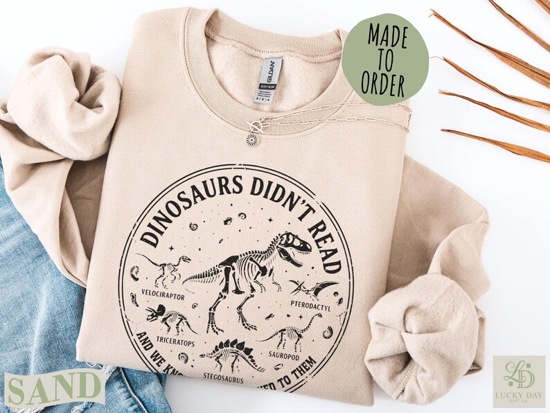 Sand sweatshirt with "Dinosaurs Didn't Read" graphic and dinosaur skeletons.