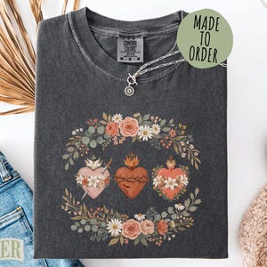 May include: A charcoal gray t-shirt featuring a graphic print of three hearts surrounded by a floral wreath. The center heart is wrapped in thorns, while the side hearts are adorned with flowers and a sword. The text MADE TO ORDER appears in a circle.