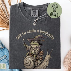 May include: A charcoal gray t-shirt featuring a graphic of a frog wearing a wizard hat and robe, riding a snail while holding a star-tipped wand. The text above the frog reads, Off to cause a kerfuffle. The shirt has a crew neck and short sleeves.