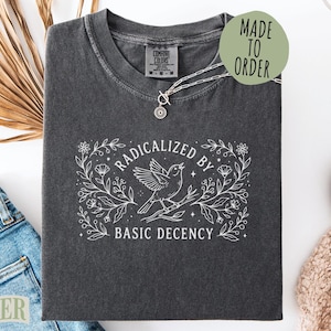 May include: Dark gray t-shirt with the text "Radicalized by Basic Decency" in a white floral design. A silver necklace with a small pendant is on the shirt. The shirt is folded on a white surface with jeans and a beige knit item.