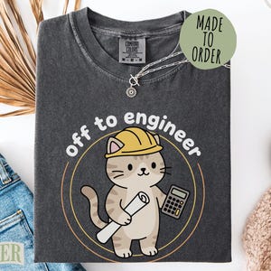 May include: A charcoal gray t-shirt featuring a graphic of a light brown tabby cat wearing a yellow hard hat. The cat holds a rolled blueprint and a calculator. The text "off to engineer" is printed in white above the cat inside a circle.