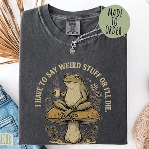 May include: A charcoal gray t-shirt featuring a graphic of a frog sitting on a mushroom while holding a steaming mug. The text "I HAVE TO SAY WEIRD STUFF OR I'LL DIE" is printed in a circular arc around the frog, surrounded by small flower illustrations.