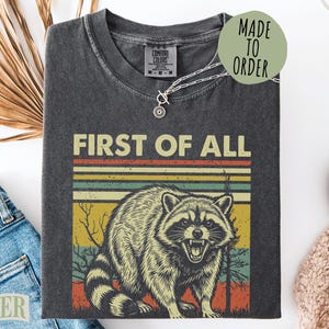 T-shirt with a vintage raccoon graphic and "First of All" text.