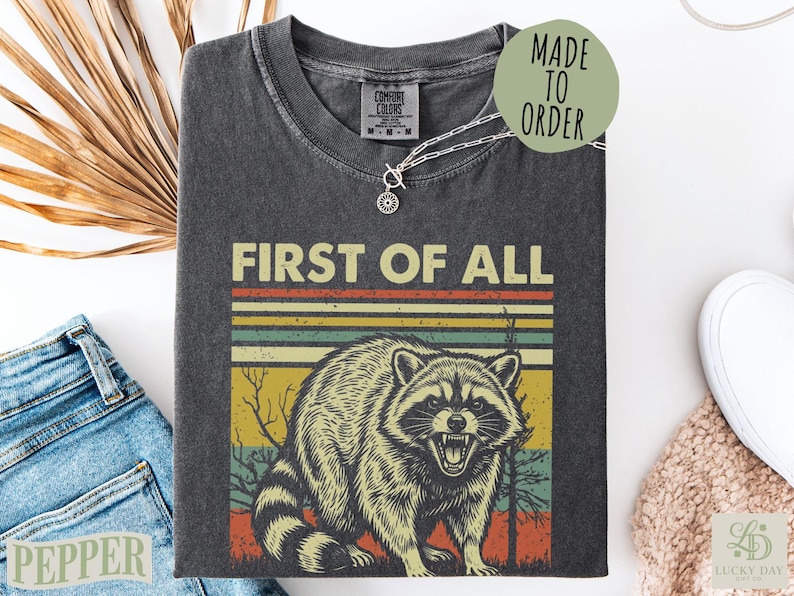 T-shirt with a vintage raccoon graphic and "First of All" text.