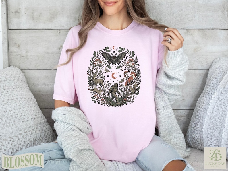 Pink t-shirt with circular forest illustration of animals and plants, featuring Bigfoot.