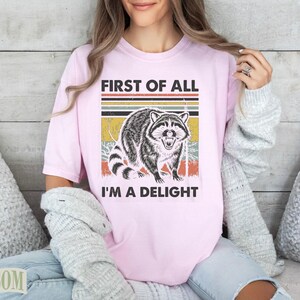 Pink t-shirt with raccoon graphic and text "First of all I'm a Delight".