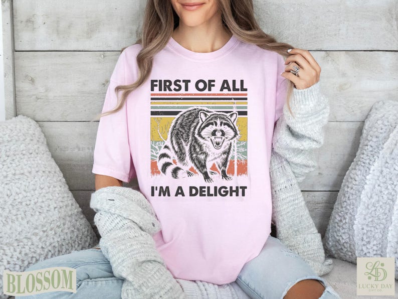 Pink t-shirt with raccoon graphic and text "First of all I'm a Delight".
