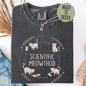 May include: A charcoal gray crew neck t-shirt featuring a graphic of four cats in a circular cycle labeled Scientific Meowthod. The design includes the words Observation, Hypothesis, Experiment, and Analysis with small paw prints and arrows.