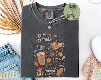 May include: A charcoal gray t-shirt featuring a graphic print with the text Cozy October, Fall Stories, Pumpkin Spice, Leaves, and Autumn Days. The design includes illustrations of a book, a coffee cup, a pumpkin spice drink, leaves, mushrooms, and a mitten.