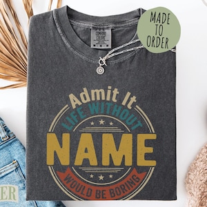 May include: A charcoal gray short-sleeve t-shirt featuring a circular graphic with the text "Admit It LIFE WITHOUT NAME WOULD BE BORING" in yellow, teal, and red. The shirt has a crew neckline and a Comfort Colors brand label at the inner collar.
